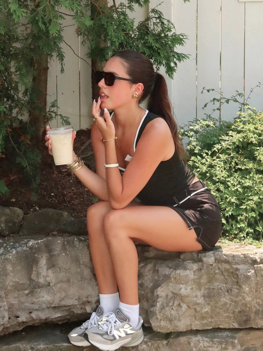 Woman sitting on a stone ledge outdoors, holding a drink and wearing sunglasses.