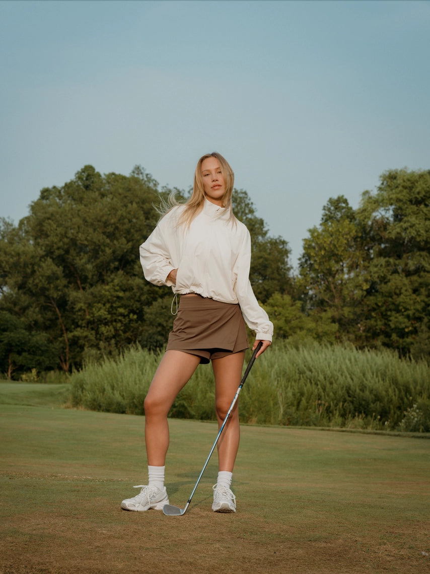 Woman holding a golf club on a golf course with trees in the background in a brown BOV skort