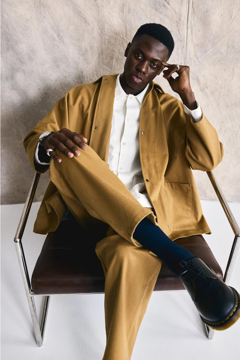 Man in a mustard jacket and pants sitting on a chair against a neutral background Devlyn van Loon 