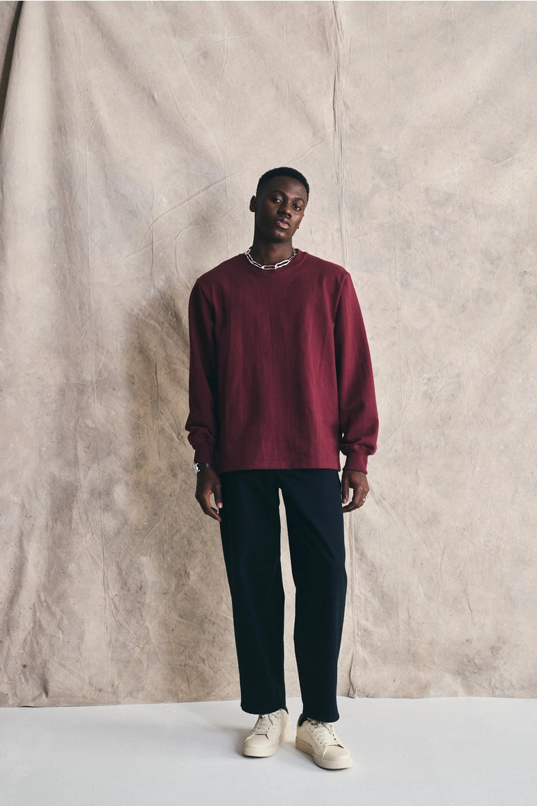 Man wearing a red sweater and black pants standing against a textured beige wall. Devlyn van Loon